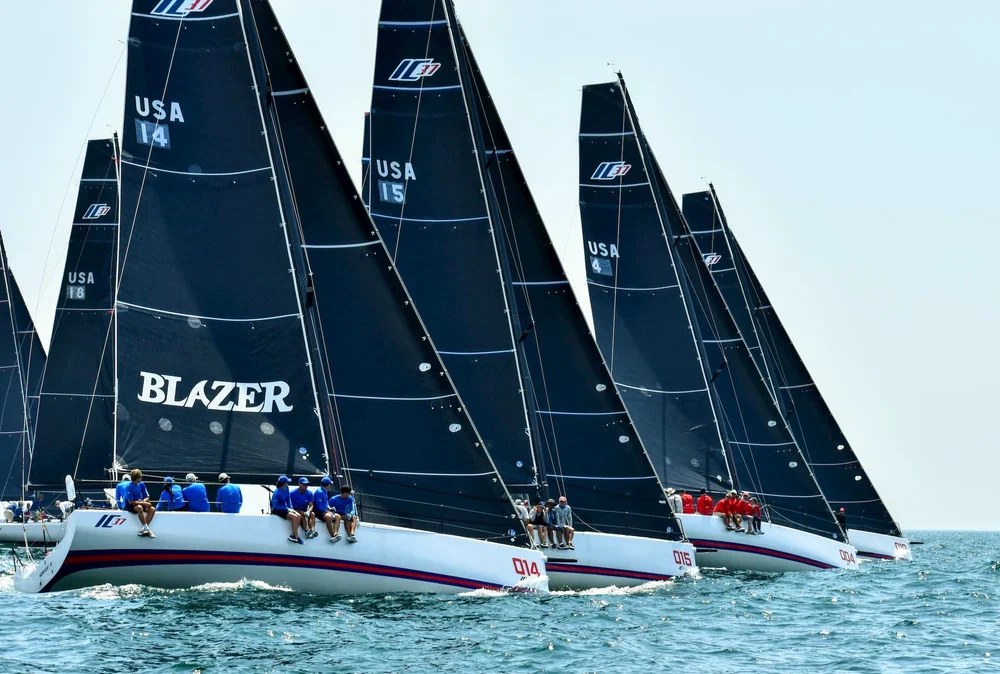 A fleet of one-design racing sailboats with dark sails, crews on the rail
