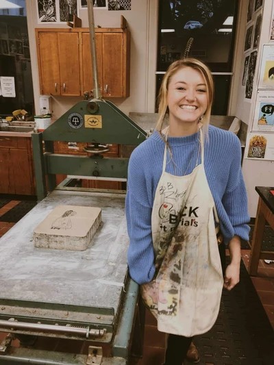 Artist Mallie Sykes smiling in a printmaking studio, wearing a paint-covered apron
